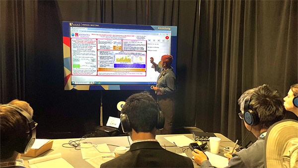 Hemali Angne, a PhD candidate in computer science at Rutgers, stands before a presentation board. They are presenting at the AAAS poster competition before the judging panel. Four judges are in the foreground of the photo, wearing headphones and looking at the poster board and presentation.