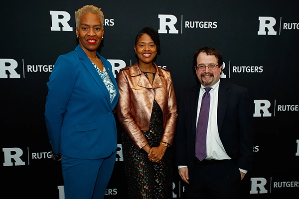Three people pose together in front of a black step-and-repeat backdrop with repeated Rutgers “R” logos. From left to right: Dr. Patti O'Brien Richardson, Associate Teaching Professor and Bloustein School Chief Community and Belonging Officer. Dr. O'Brien Richardson is a Black woman in a blue suit, smiling with her hands at her sides. Next is Dr. Chris T. Pernell, director of the NAACP Center for Health Equity. Dr. Pernell is a Black woman in a gold jacket and patterned dress standing with her hands clasped in front of her. The third person is Bloustein School Dean Stuart Shapiro, a white a man in a dark suit with a tie. The jacket is open and his hands are by his side, and he smiles slightly. All three face the camera.