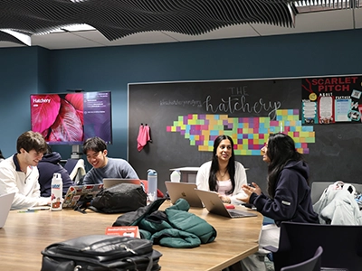 A group of four diverse students sit at a table talking to each other. There are backpacks, clothing, laptops and water bottles on the table in front of them. The background is a black board and colorful tape flags and posters.