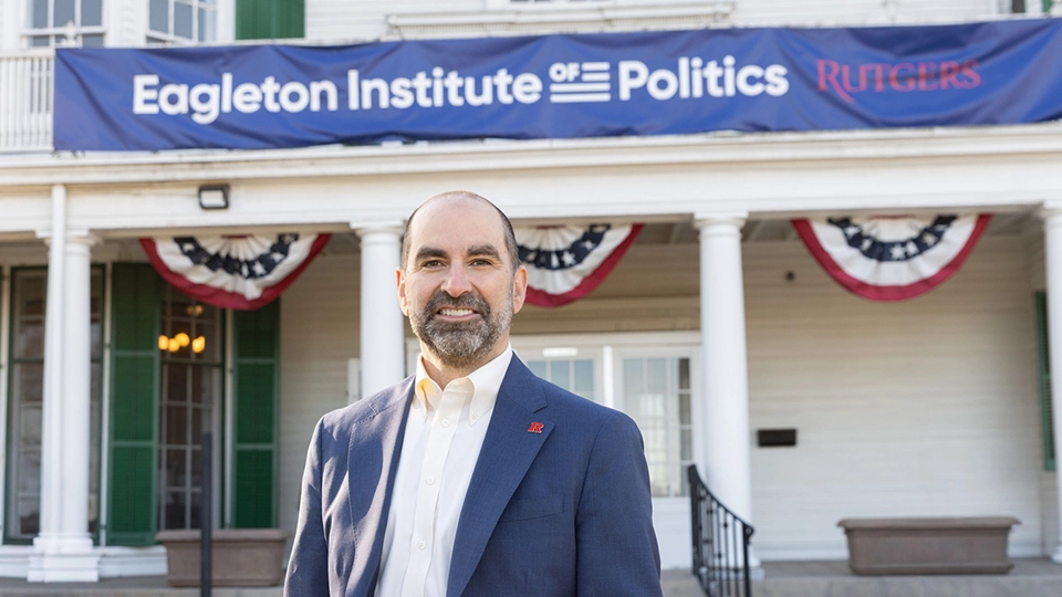 Nick Longo, a smiling male with a beard wearing a white shirt and blue jacket with a a red Rutgers "R" lapel pin, stands in front of a white house with "Eagleton Institute of Politics at Rutgers" on a blue banner.